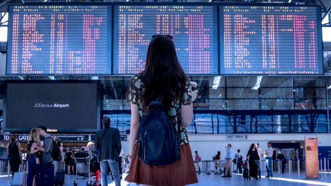 Girl standing back facing the camera in an airport toward the flight information display system