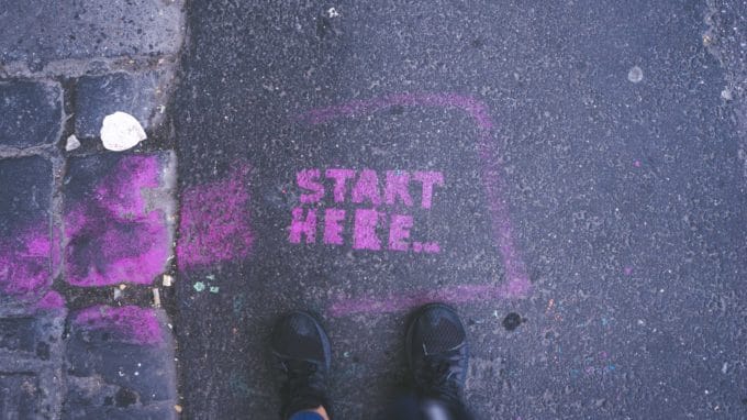 "Start here" spraypainted in pink on the road with a person's feet at the "start here" wording